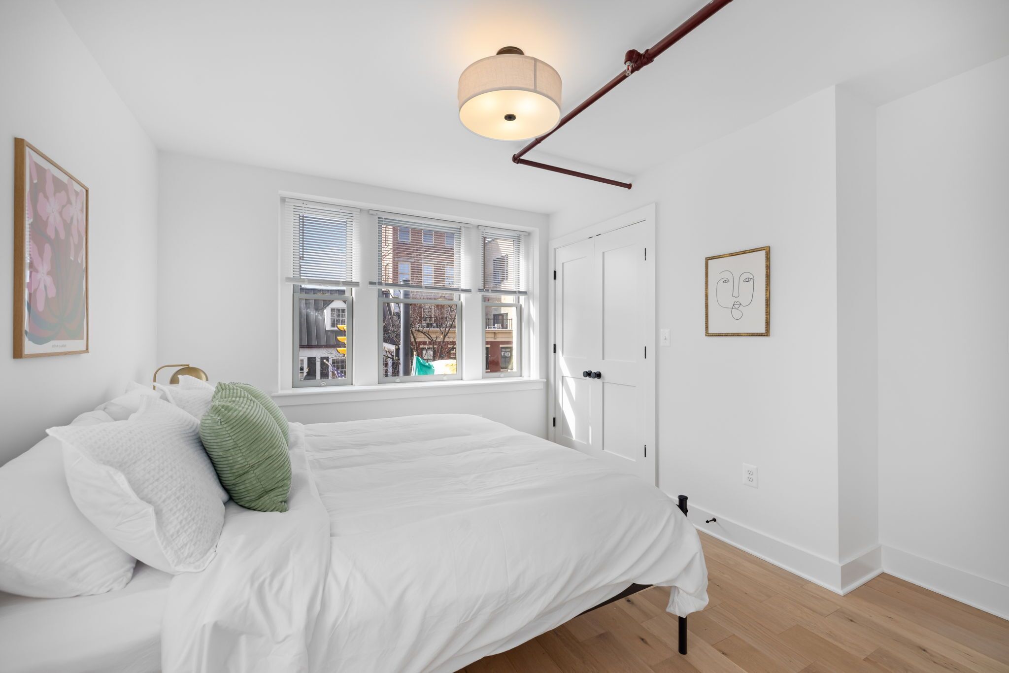 Une chambre à coucher avec un lit, une fenêtre avec des stores, une porte et un luminaire fixé au mur.