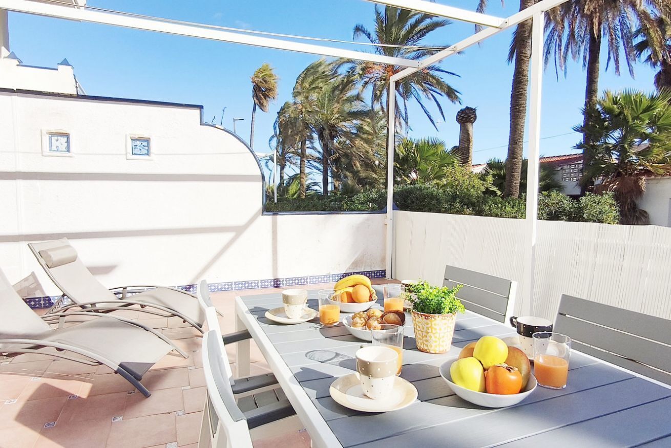 A balcony with a table set for breakfast, including fruits, a plant, and a view of palm trees.