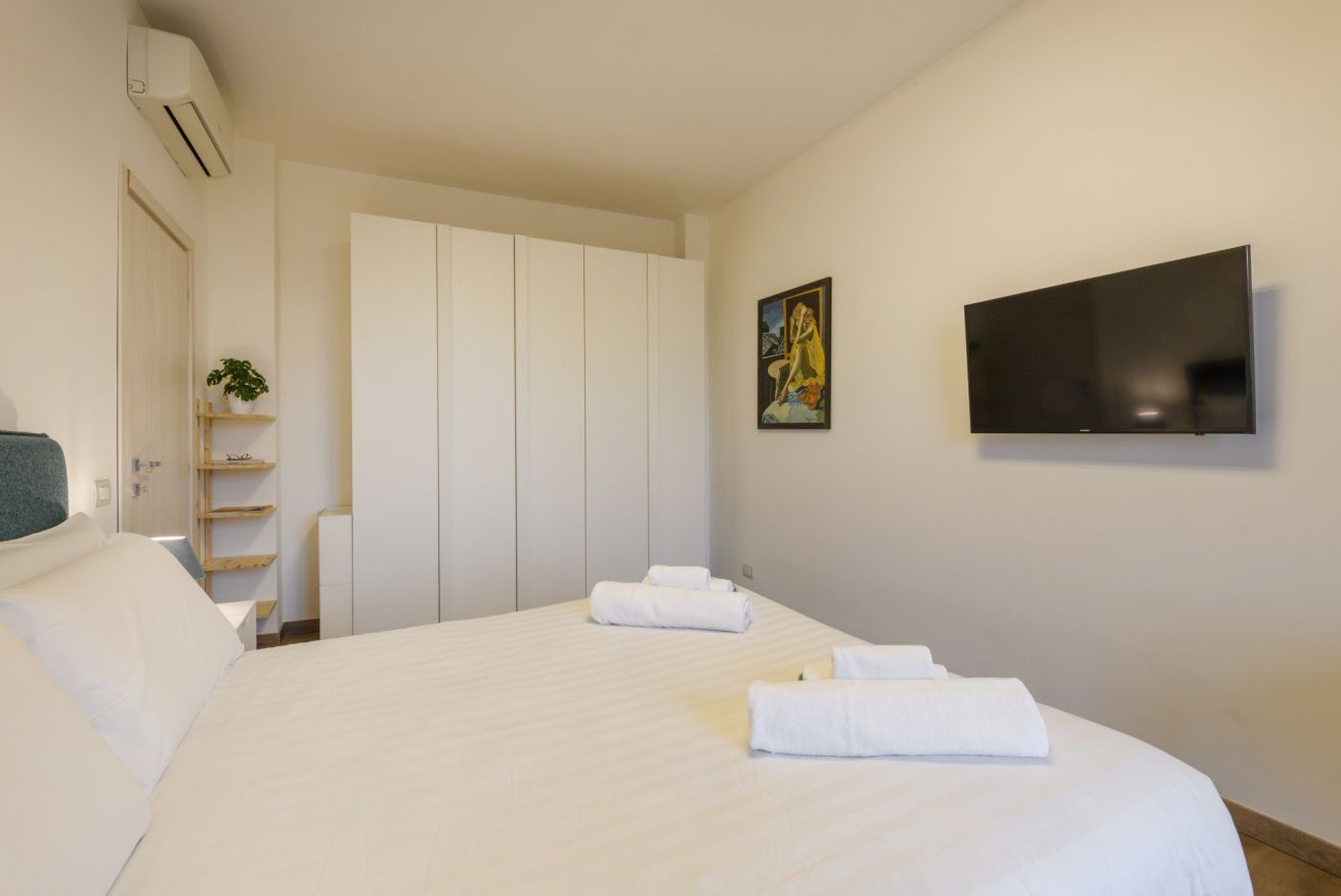 A hotel room with a bed, a flat-screen TV, a painting, a wardrobe, and a plant.