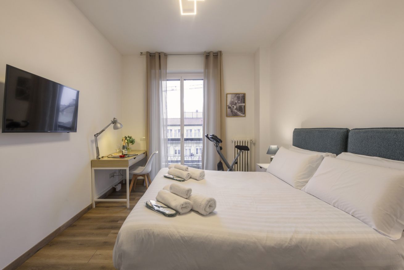 A hotel room with a bed, a desk with a lamp, a TV, and a window with curtains.