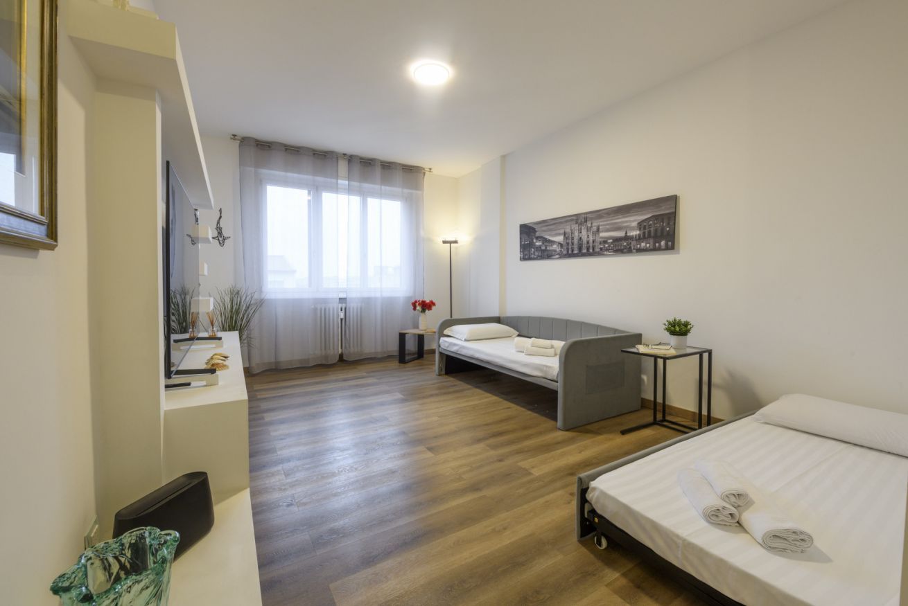 A modern hotel room with a bed, a desk, a chair, a small table with a plant, and a window with curtains.