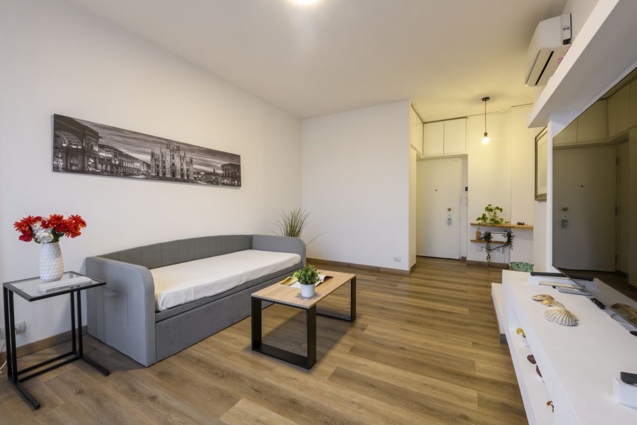 A modern living room with a grey sofa, a small table with a vase of flowers, and a painting on the wall.