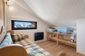 Une chambre mansardée chaleureuse, avec un lit, une étagère remplie de livres et un téléviseur, ainsi qu’une fenêtre donnant sur les montagnes.