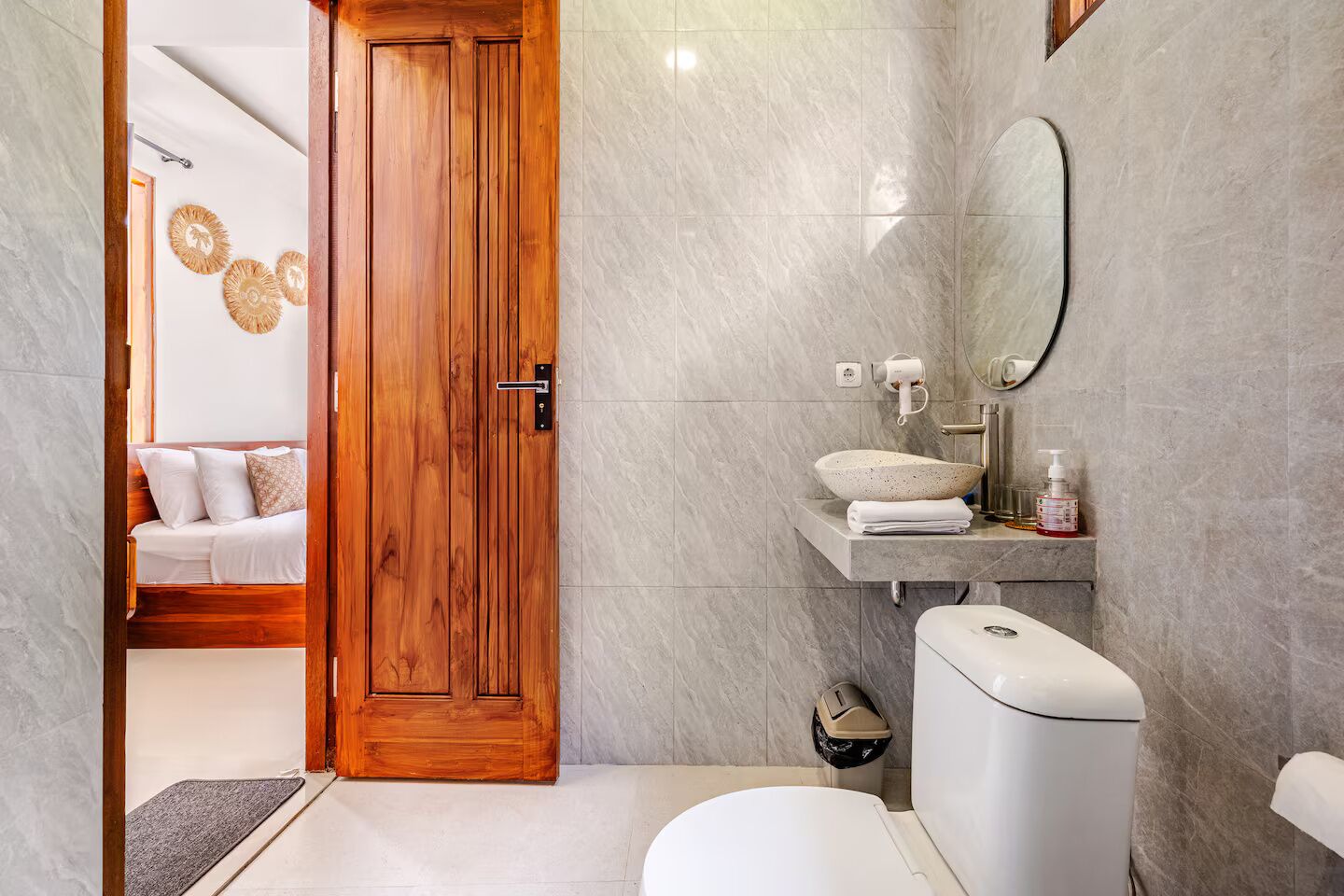 A bathroom with a toilet, sink, mirror, and a wooden door leading to a bedroom.