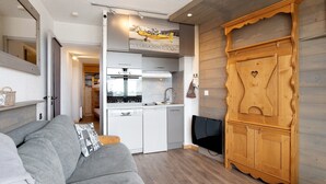 A compact living space with a kitchenette, a grey sofa, and a wooden cabinet. - Cozy Ski-In/Ski-Out Studio 26 m² – Alpe d’Huez (Huez)