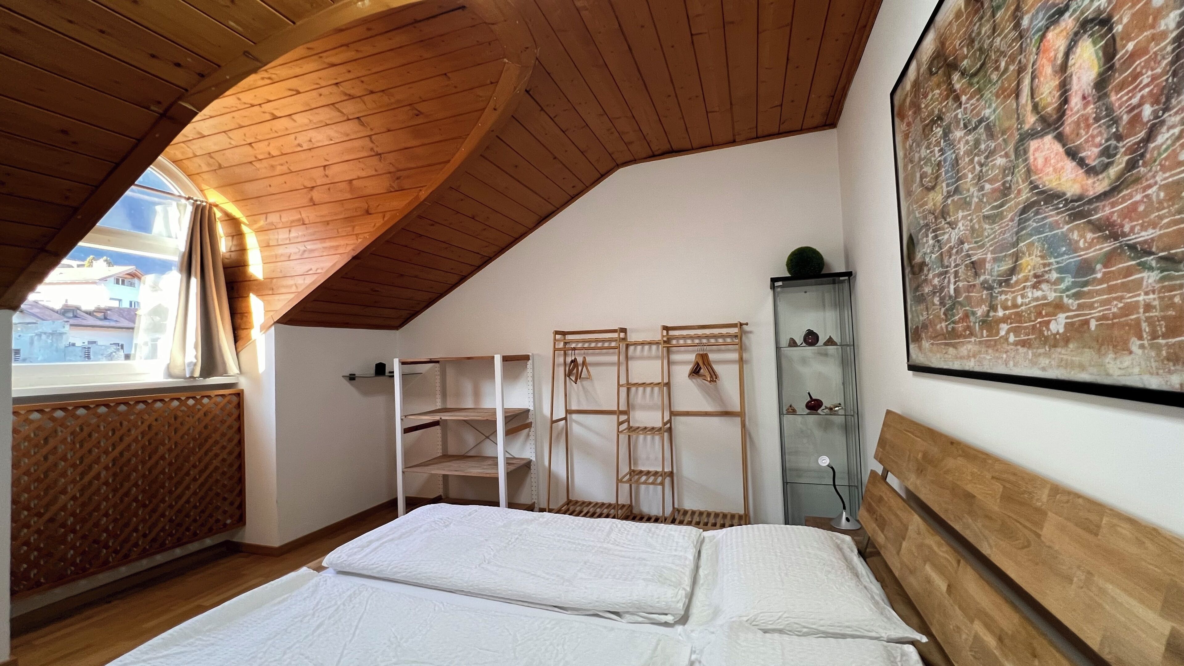 Une chambre avec un plafond en pente, des poutres en bois, un lit, une armoire et une fenêtre donnant sur des immeubles.