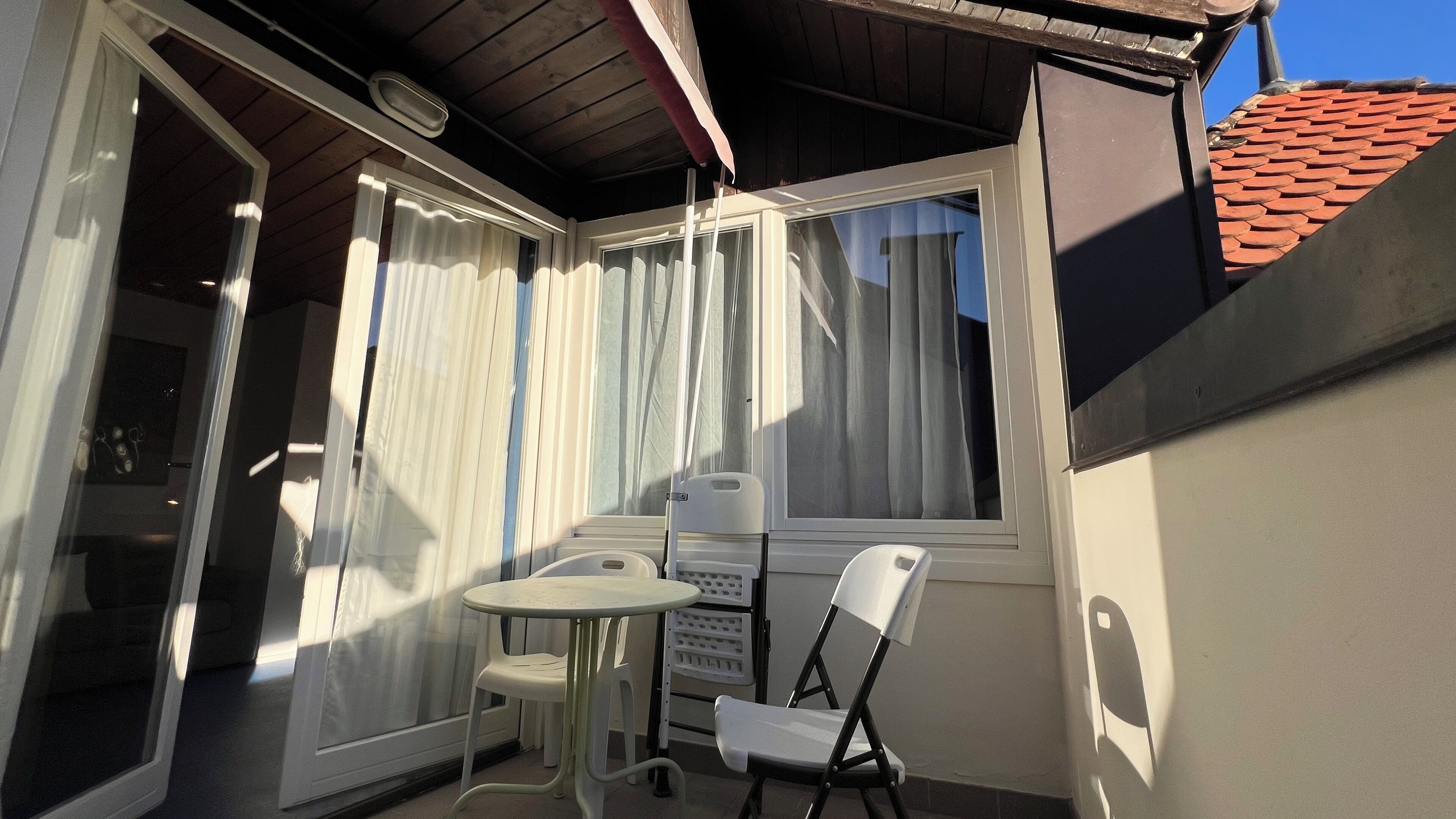 Ein kleiner Balkon mit Tisch und Stühlen, ein Fenster mit Vorhängen und ein Ziegeldach.