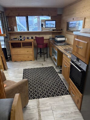A compact kitchen with wooden cabinets, a built-in oven, a dining area with a red chair, and a large window. - Charming Apartment for 8 – Ski-In/Ski-Out at Alpe d'Huez (Huez)