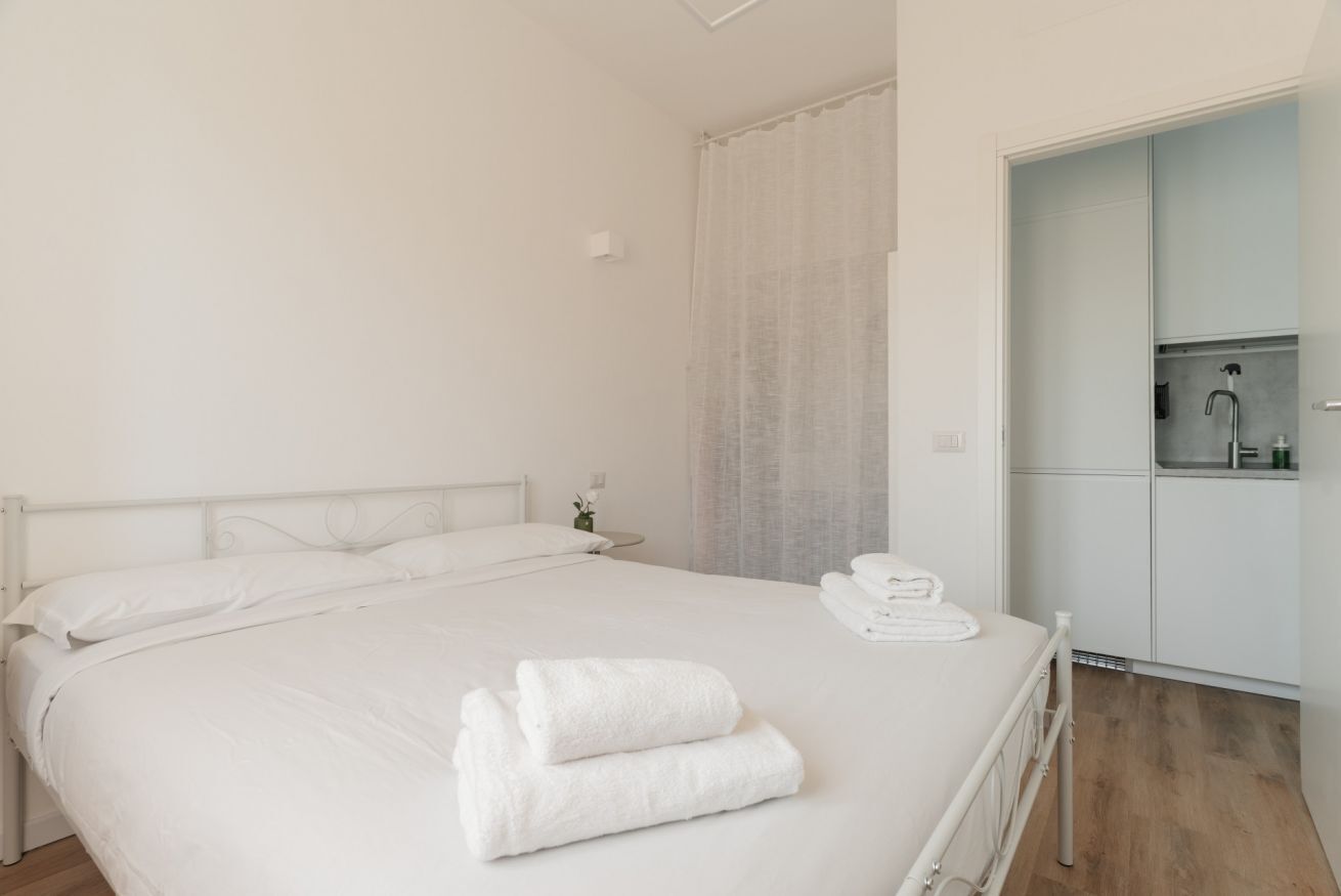 Una cama bien hecha con ropa blanca y toallas, una mesita de noche con un florero, y un armario con espejo.