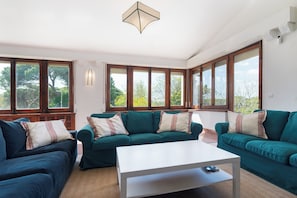A living room with two green sofas, a white coffee table, and large windows with a view of trees. - Luxurious finca with swimming pool near the beach. (El Puerto de Santa María)