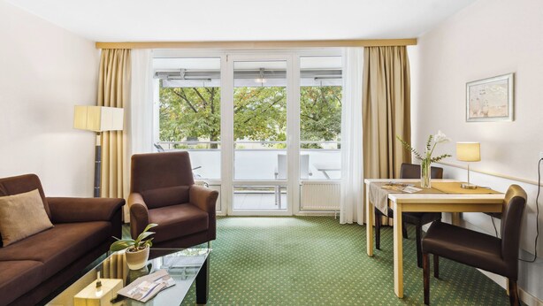 A hotel room with a green carpet, a sofa, a chair, a dining table, and a window with curtains. - Cozy and quiet vacation apartment in the center of Bad Füssing (Bad Füssing)