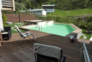 Piscina com deck de madeira, espreguiçadeira e mesa pequena.