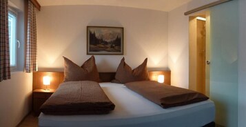 Uma cama de casal com duas almofadas, uma mesa de cabeceira com abajur, um quadro na parede e uma porta à direita.