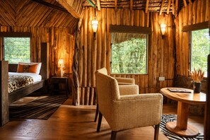 Una habitación rústica de madera con una cama, una silla de mimbre, una mesa y una ventana con vistas a la vegetación.
