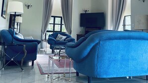 Un salon avec un canapé bleu, des fauteuils, un téléviseur et un lustre.