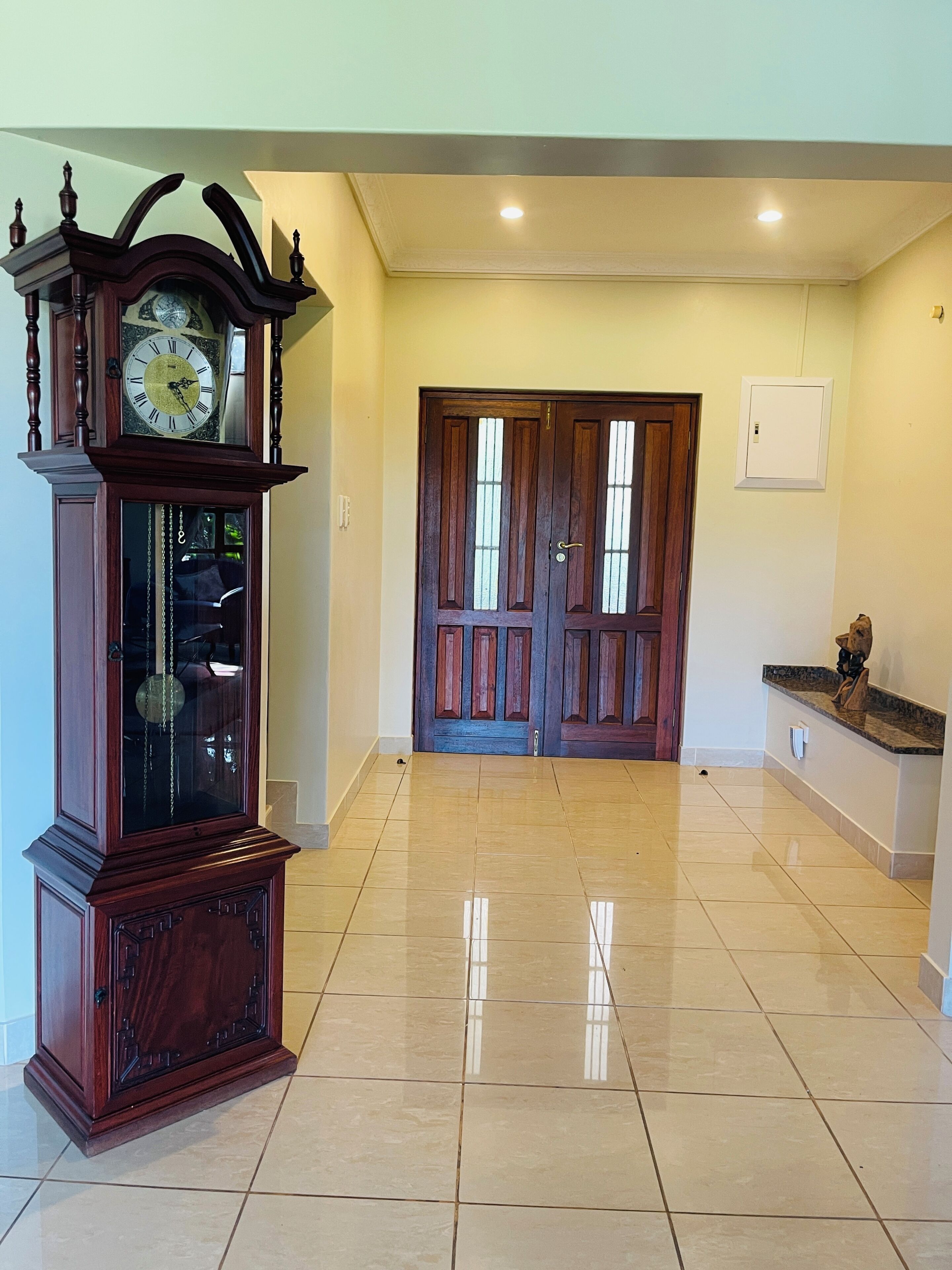 Un couloir doté d’un plancher en bois, d’une grande horloge murale, de portes à deux vantaux et d’un petit comptoir sur lequel est posé un objet décoratif.