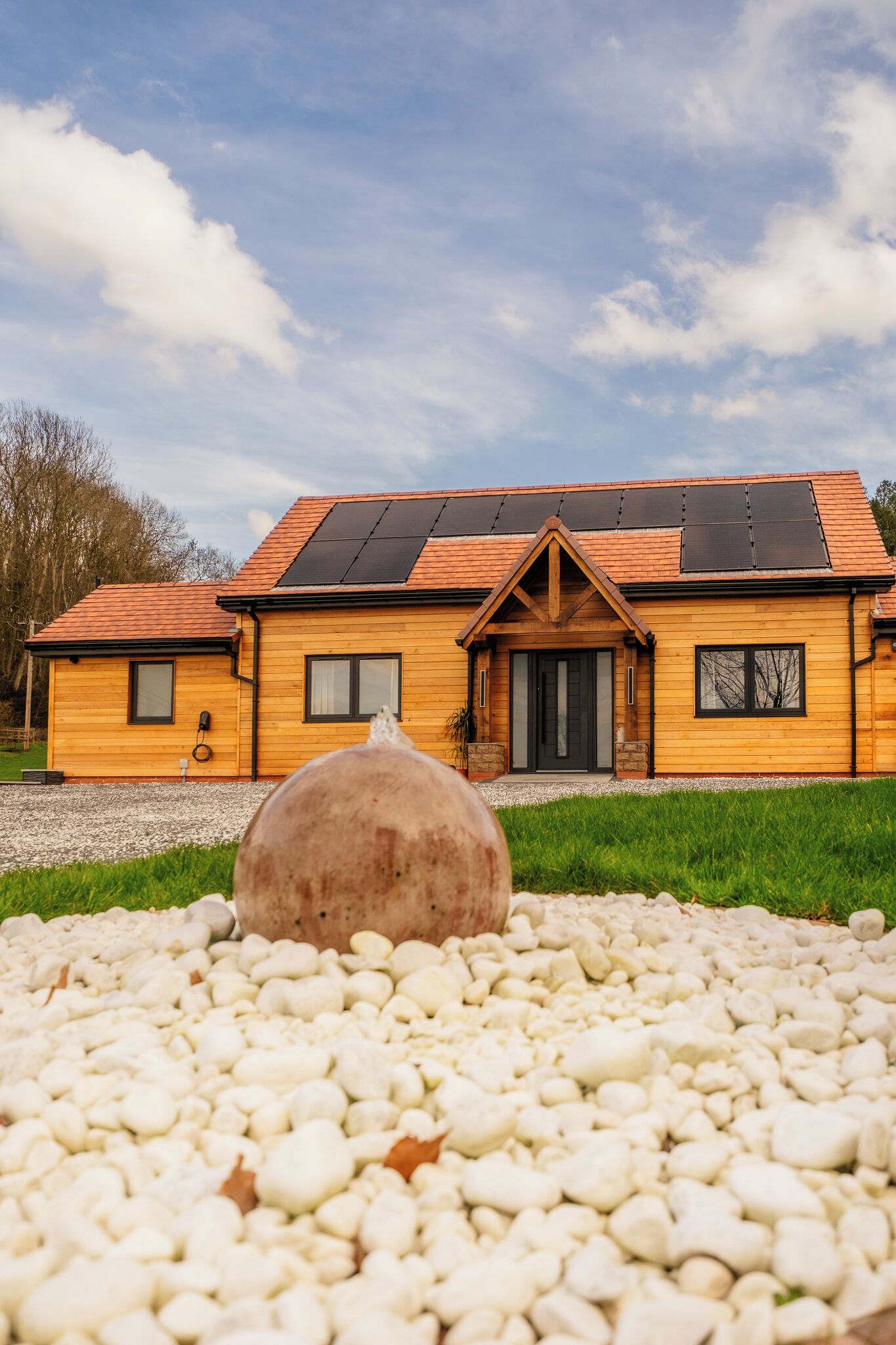 A wooden house with a sloped roof and solar panels, a front door, and a driveway with white pebbles.