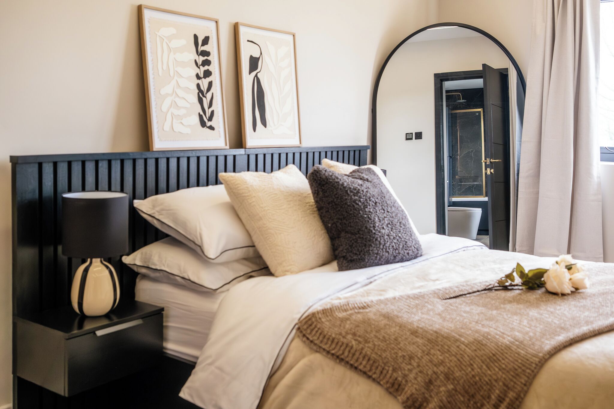 Quarto de hotel com uma cama, uma mesinha de cabeceira com abajur, um espelho e duas obras de arte emolduradas na parede.