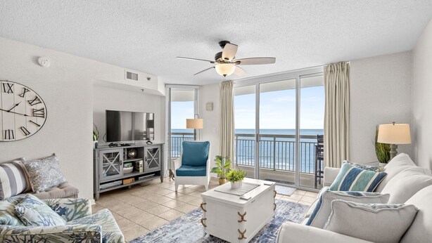 A living room with a large clock, a ceiling fan, a TV, a blue chair, and a view of the ocean. - 20% off 3-7 thru Dec. Oceanfront 2BR 2BA. Crescent Shores. Views!! Amenities!! (North Myrtle Beach)