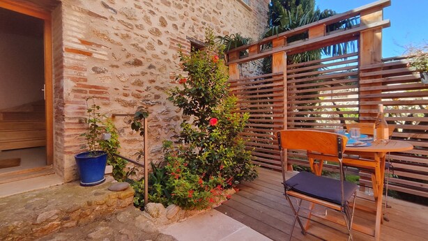 Une terrasse en bois avec une chaise et une table, un mur en pierre et une pergola en bois recouverte de plantes grimpantes.
