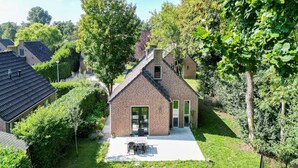 A brick house with a sloped roof, a front door, and a small patio with a table and chairs. - Woodland Bungalow Retreat for 4 (Ewijk)