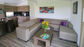 A modern living room with a sectional sofa, a dining area with chairs, a coffee table with books, and a kitchen in the background. - Chalet With 3 Bedrooms and Infrared Sauna (West-Graftdijk)
