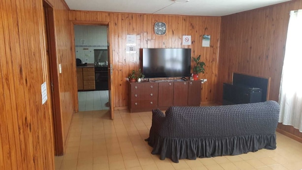 A living room with a TV, wooden walls, a sofa, and a kitchen in the background.