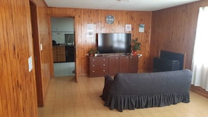 A living room with a TV, wooden walls, a sofa, and a kitchen in the background.
