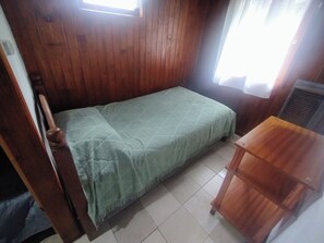 A room with a wooden wall, a bed with a green blanket, a small wooden table, and a window with curtains.