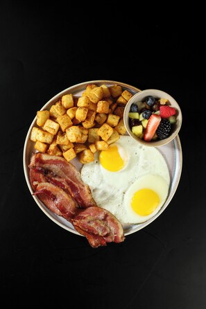 Un plato de desayuno con huevos, tocino, papas y un bol de fruta.