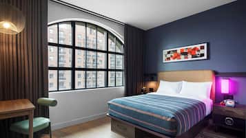 A hotel room with a large window, a bed with patterned bedding, a desk with a chair, and a nightstand with a lamp and phone.