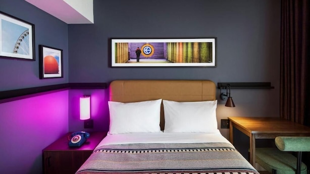 Un cuarto de hotel con cama, mesita de noche, lámpara, un cuadro en la pared y vista a un edificio.