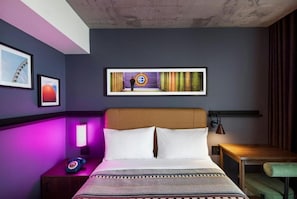 A hotel room with a bed, a nightstand, a lamp, a framed picture on the wall, and a view of a building outside. - Moxy Ottawa Downtown (OTTAWA)