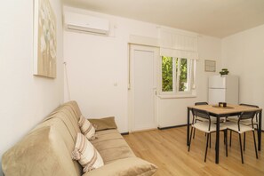 A room with a beige sofa, a wooden dining table with chairs, a white door, and a window with a view of greenery. - Stylish vacation apartment in Dramalj - beach just 250 m away (Dramalj)