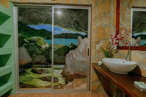 Une salle de bain avec un mur en pierre, une grande fenêtre offrant une vue panoramique, un lavabo blanc et un miroir.