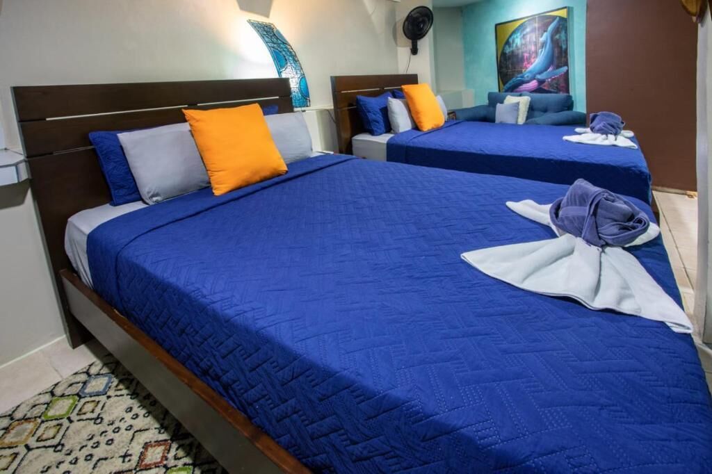 A hotel room with two beds, each with blue bedding and orange pillows. There is a fan on the wall and artwork on the wall.