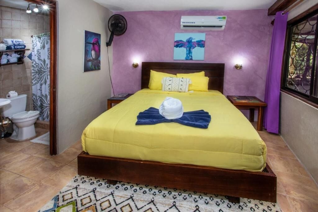 A bedroom with a bed, bedside tables, a fan, a shower curtain, a toilet, and a window with a view.