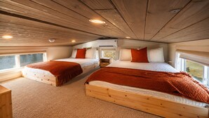 Interior de cabana com duas camas de madeira, uma janela e teto com vigas de madeira.