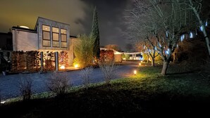 A modern building with large windows, a paved driveway, and a well-lit garden area with trees and lights.