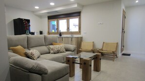 Una sala de estar con sofá, dos sillones, una mesa de centro de vidrio, una zona de cocina con ventana y una puerta.