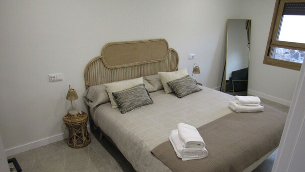 Un dormitorio con cama, almohadas, lámpara de noche, espejo y ventana.