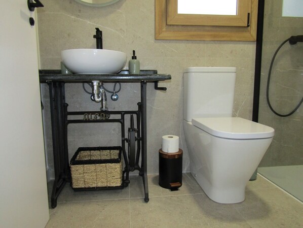 A bathroom with a white toilet, a sink with a basin, a toilet paper holder, and a small trash bin.