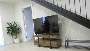 Una escalera de diseño moderno, un televisor de pantalla plana sobre un soporte de madera, una planta en maceta y un perchero metálico.