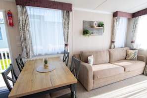 A cozy living room with a wooden table, beige sofa, and a wall-mounted shelf with plants. - SBL63 Parkdean Resorts Camber Sands (Camber)