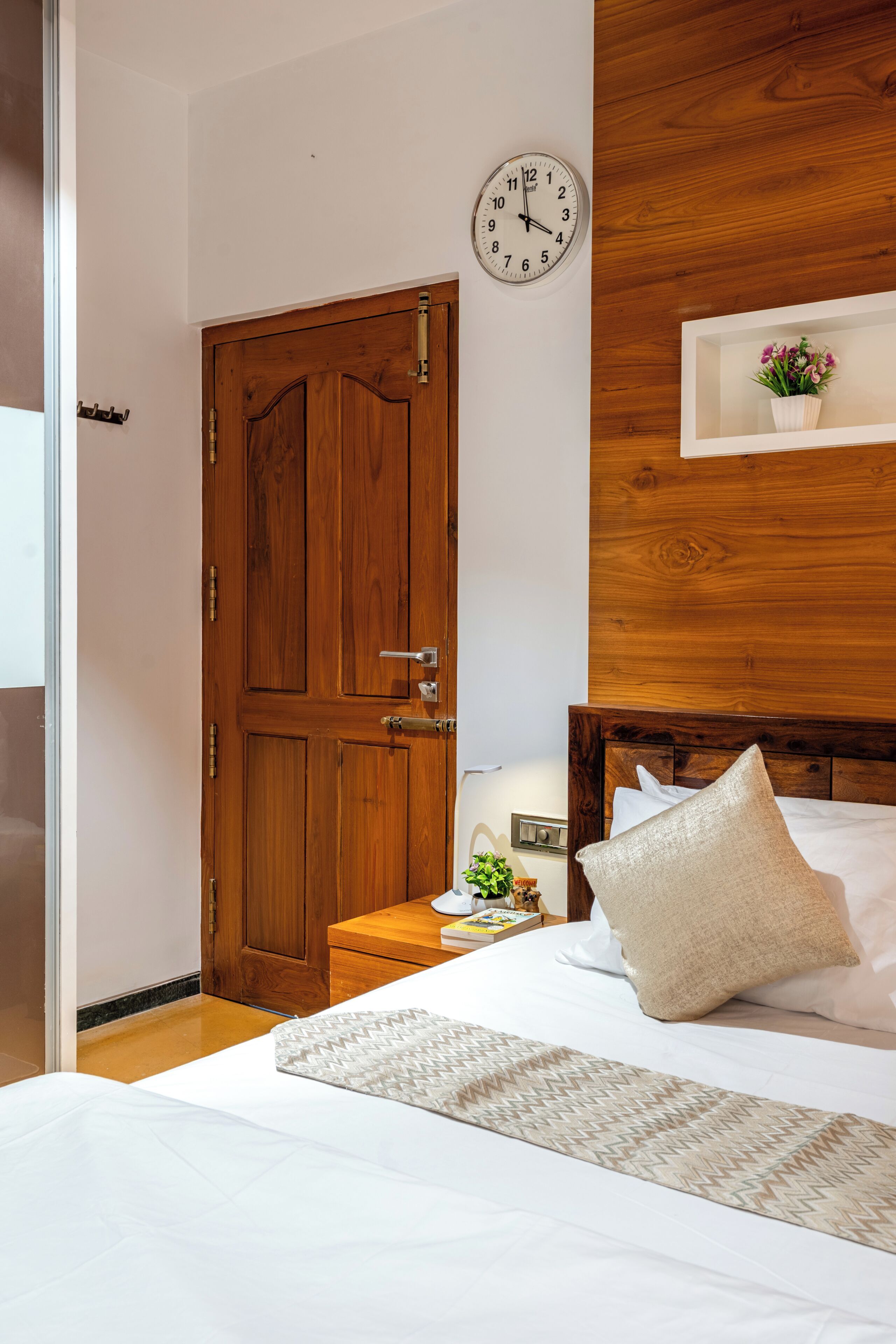 A bedroom with a wooden door, a bedside table with a lamp, a clock on the wall, and a shelf with a plant.