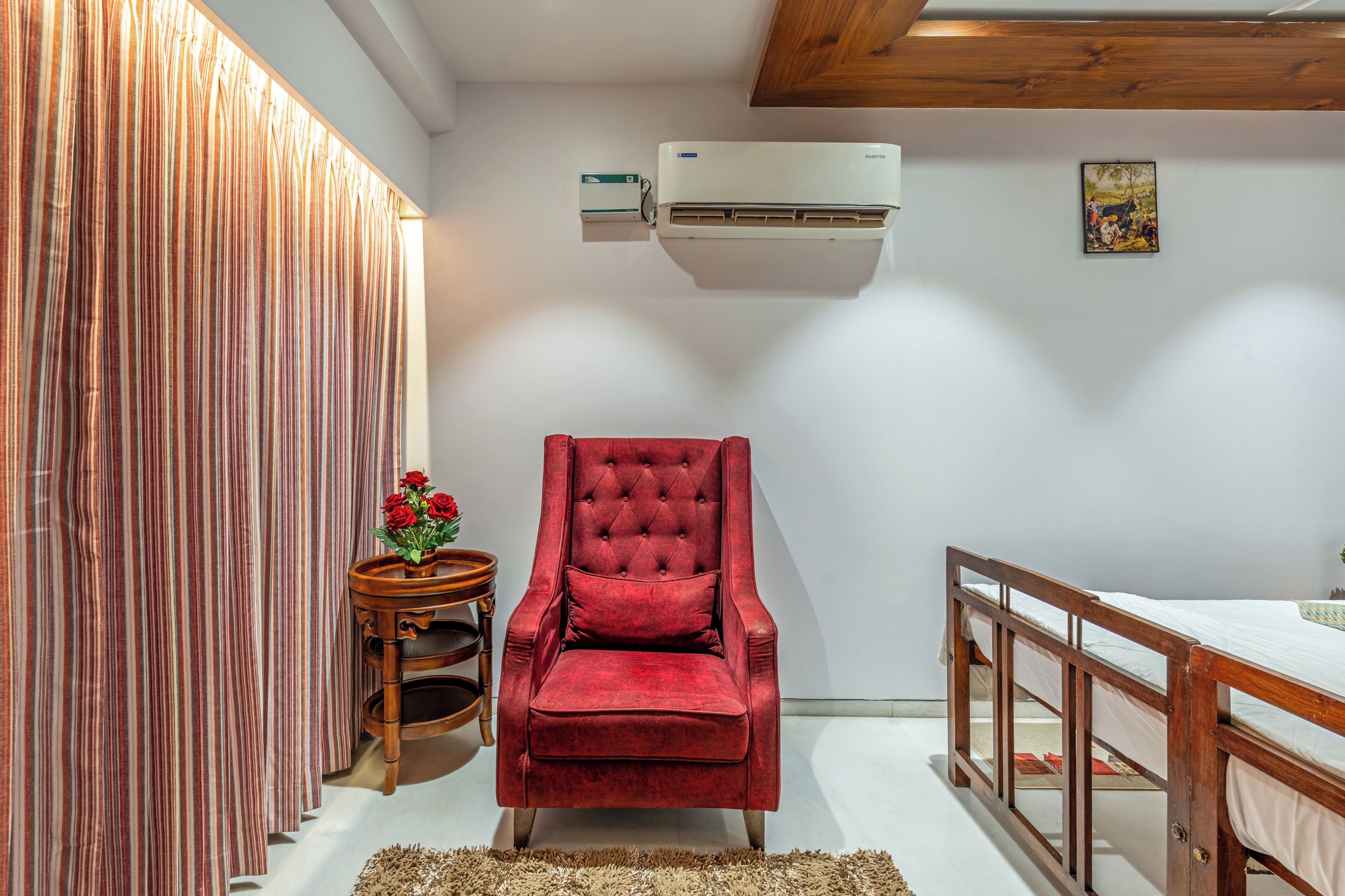 Una habitación con una butaca de terciopelo rojo, una mesita de madera con un florero y una unidad de aire acondicionado blanca, además de una viga de techo de madera.
