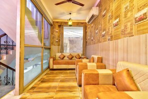 A hotel room with a wooden floor, a sofa set, a ceiling fan, and a large window with curtains. - Airport Hotel Annexe (New Delhi)