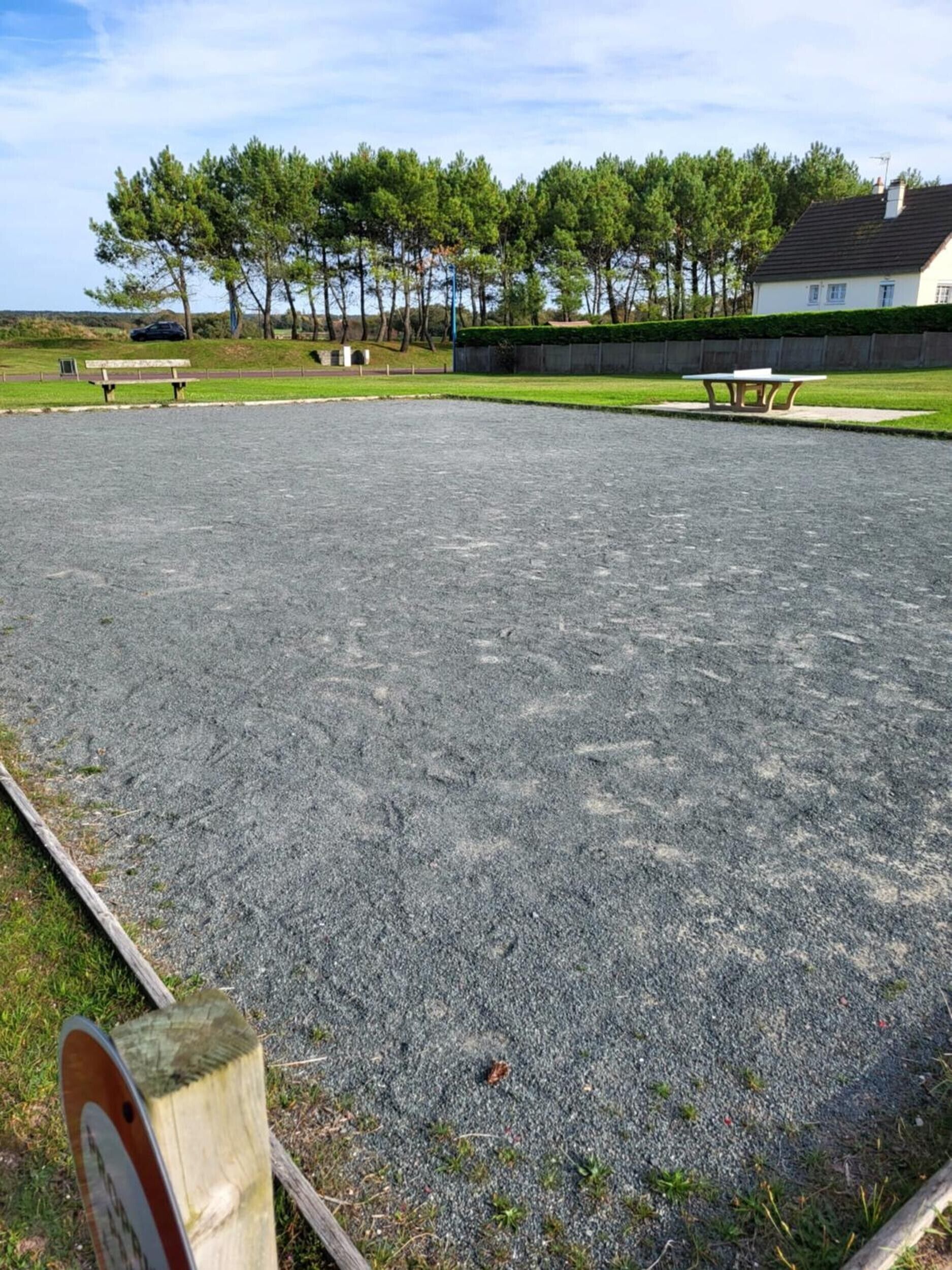 A gravel sports court with benches and a picnic table, surrounded by trees and a building.