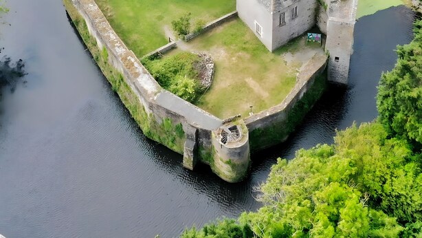 Un castillo medieval con un foso, rodeado de vegetación y un río.