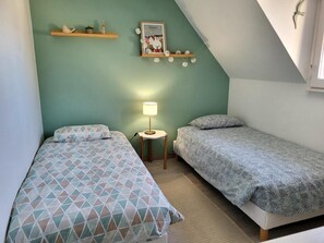 A room with two beds, a small table with a lamp, a shelf with decorative items, and a framed picture on the wall. - Beach House 'Le Cocon Pirouais' with Private Terrace, Garden, and Wi-Fi (Pirou)